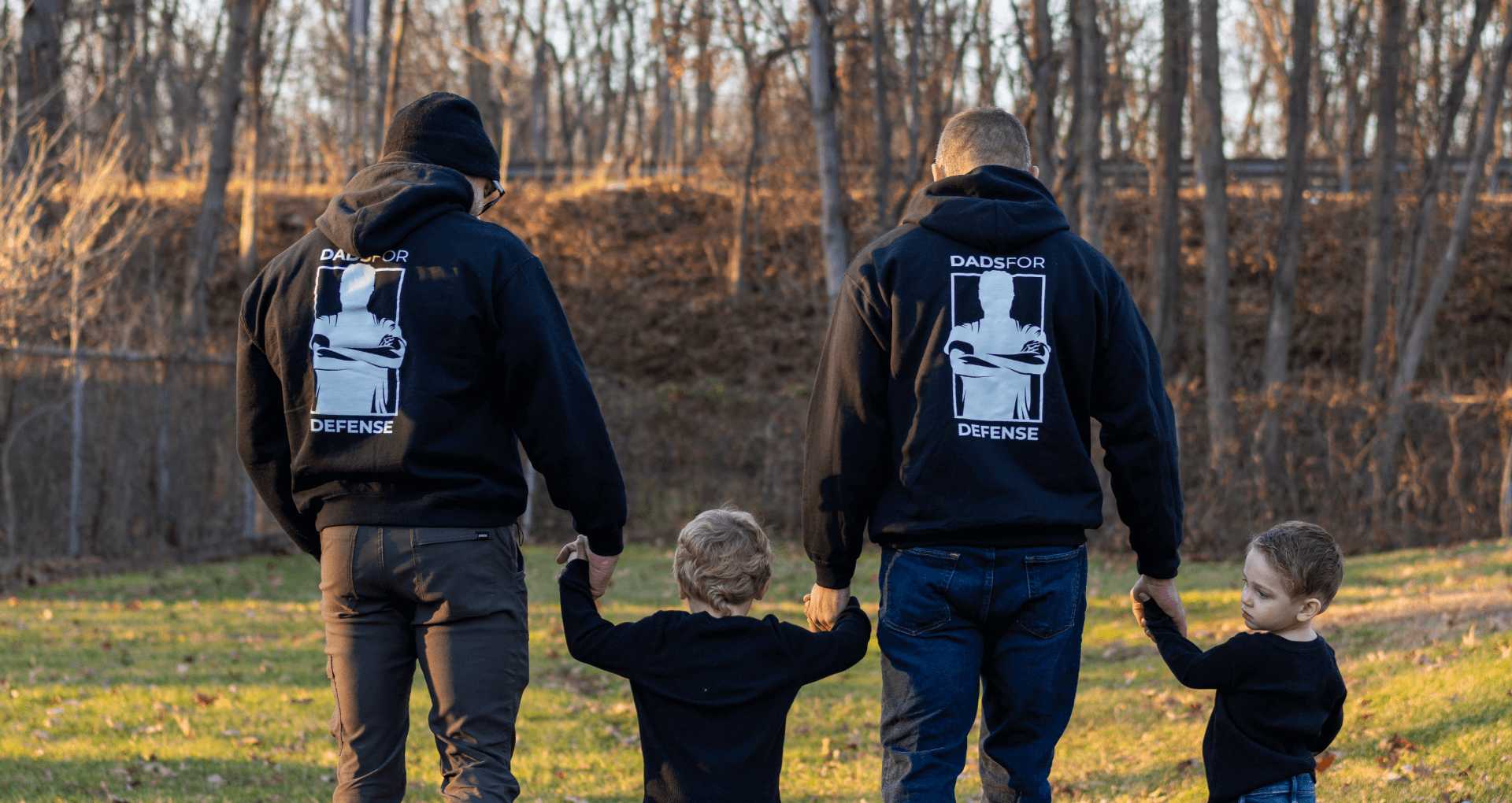 Two men and two children walking outdoors, wearing matching "dads for defense" hoodies.